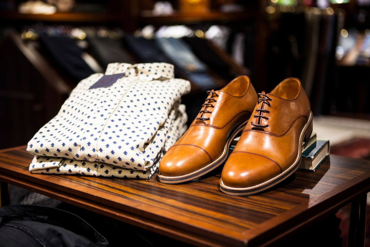 services-02 Elegant brown leather shoes and printed shirt displayed on a wooden table in a stylish clothing store.
